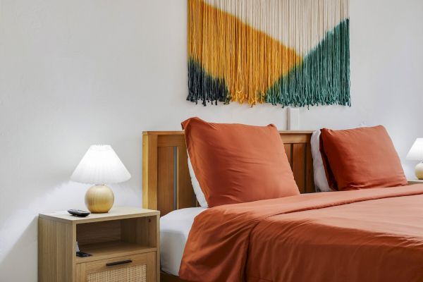 A bed with orange bedding and two wooden headboards, a wooden nightstand with a lamp, and a macrame wall hanging.