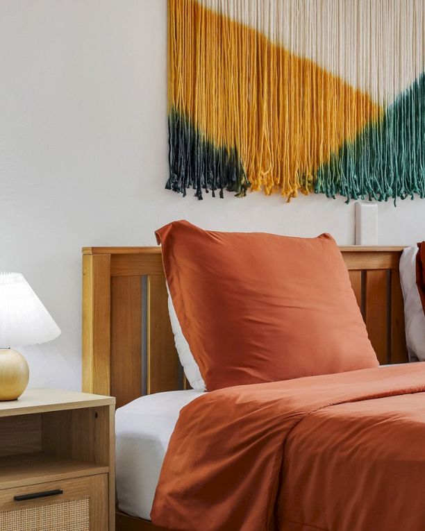 A cozy bedroom with a double bed, orange bedding, bedside lamps, and a multicolor fringed wall textile above it.