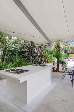 An outdoor patio with a covered shade sail, a built-in grill island, patio chairs, and lush tropical plants.