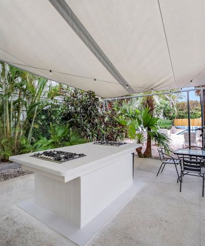 An outdoor patio with a covered shade sail, a built-in grill island, patio chairs, and lush tropical plants.