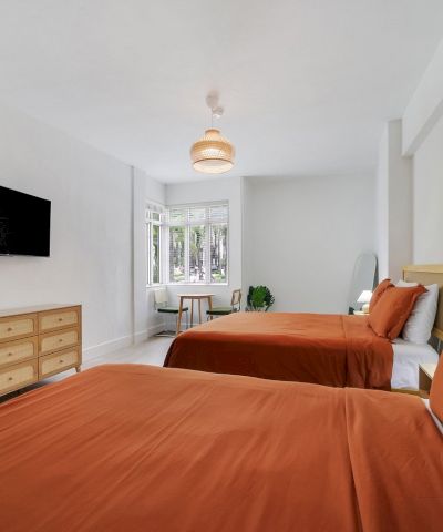 A bright modern bedroom with twin beds, orange linens, a TV, a dresser, and a woven pendant light above.