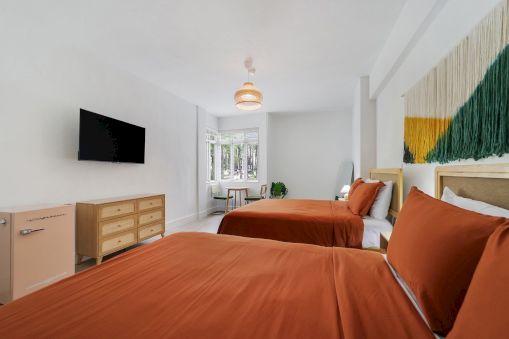 A bright modern bedroom with twin beds, orange linens, a TV, a dresser, and a woven pendant light above.