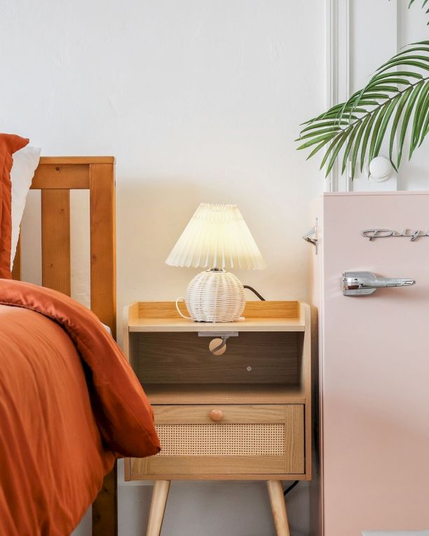 A modern bedroom corner with orange bed linens, a bedside cabinet, lamp, and a potted palm bringing fresh greenery indoors.