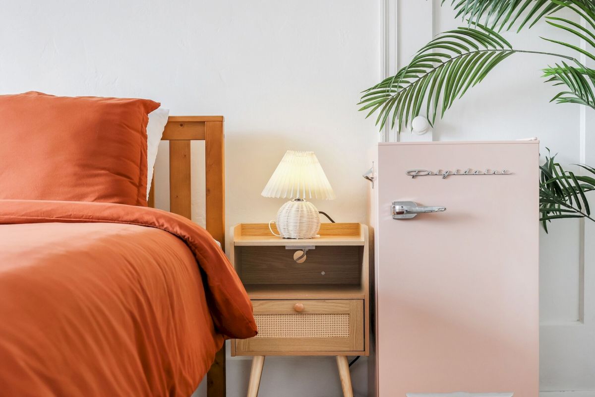 A hotel room with an orange bed, wooden nightstand, mini fridge, and a lamp.