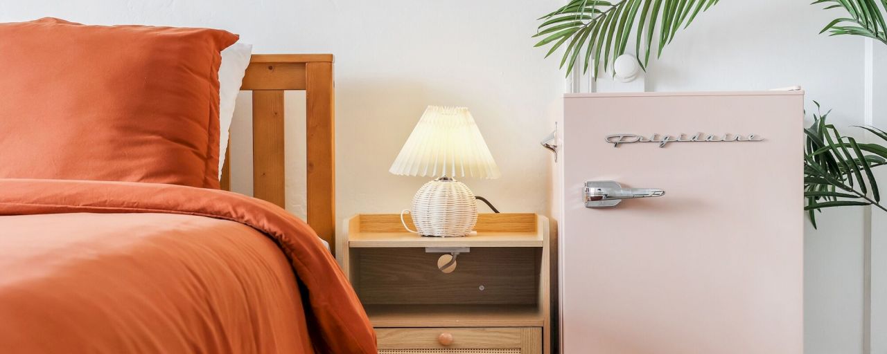 A neatly made bed with orange bedding next to a wooden nightstand with a wicker lamp, and a small pink refrigerator beside it.