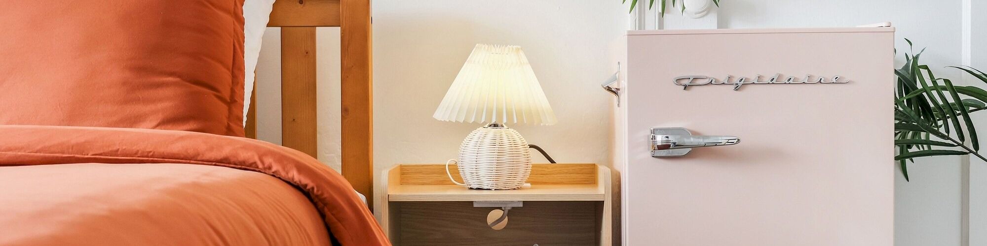 A bright bedroom corner with an orange bed, wooden nightstand, small lamp, white cabinet, and large green palm leaves.