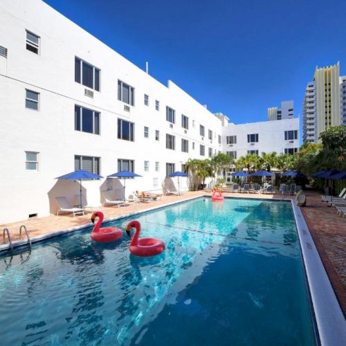 Outdoor hotel pool with blue umbrellas and pink flamingo floats, beside white buildings under a clear blue sky.