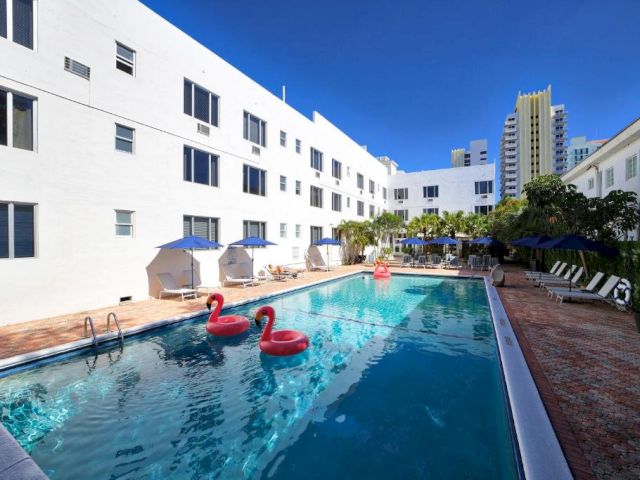 A rectangular pool with red floaties and blue umbrellas in front of a white building.