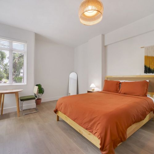 A bright modern bedroom with an orange bed, bedside tables and lamps, a window with blinds, and a woven pendant light.