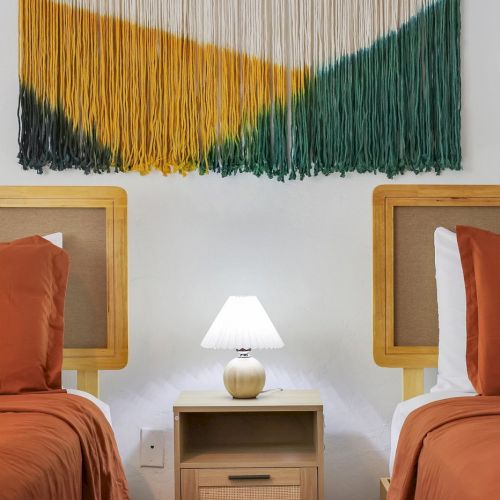 A bright hotel room with two orange beds, a bedside lamp on a wooden table, and a green-yellow fringe wall hanging.