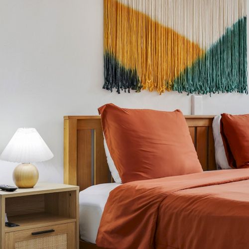 A modern bedroom with a bed made of orange bedding, lamps, and a colorful striped wall tapestry above the headboard.