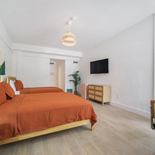 A modern bedroom with a large orange bed, white walls, TV, dresser, plant, and a small dining table by a window.