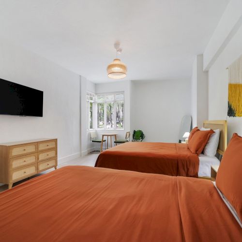 A bright modern bedroom with two beds, orange bedding, a wall TV, and a woven pendant ceiling light.