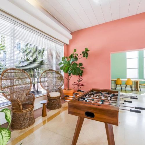 An indoor game room with a foosball table, wicker chairs, potted plants, and large windows with curtains.
