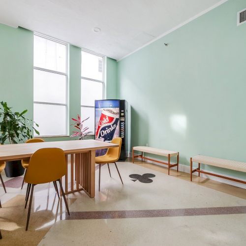 A bright office break room with mint walls, large windows, a wooden table with yellow chairs, a drink vending machine, and a bench.