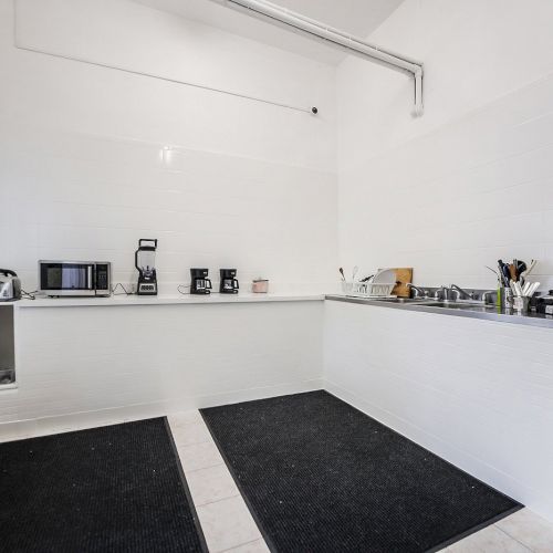 A clean commercial kitchen with white tile walls, countertop appliances, coffee makers, a sink, and metal utensil racks.