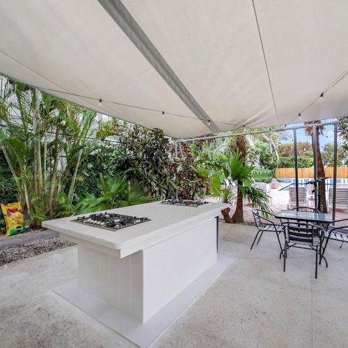 An outdoor patio garden with a large built-in BBQ grill, metal dining chairs, and string lights under a white canopy.