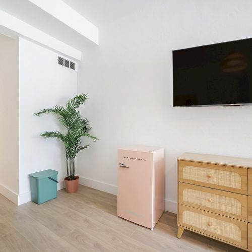 A bright living room with a wall-mounted TV, tall potted plant, and pastel storage cabinet and drawers.