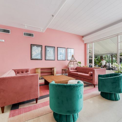 Modern lounge with pink walls, velvet couches, green chairs, large windows, and framed art.