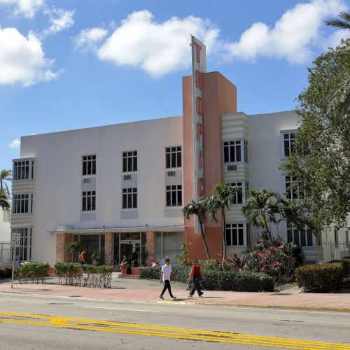A white modern campus building with a tall red vertical sign, palm trees, and people walking outside under a blue sky.