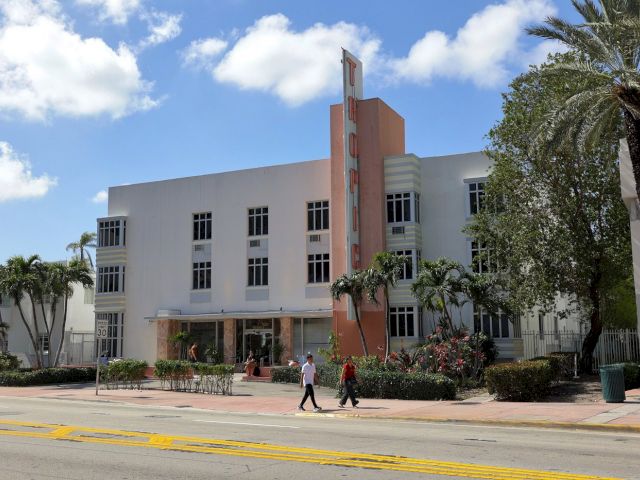 A wide exterior view of a multi-story building with art deco style, surrounded by palm trees and greenery, under a blue sky with clouds.