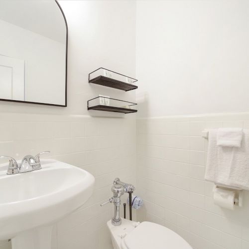 Modern white bathroom with a sink, toilet, chrome faucet, folded towels, and wall shelves under a large mirror, clean.