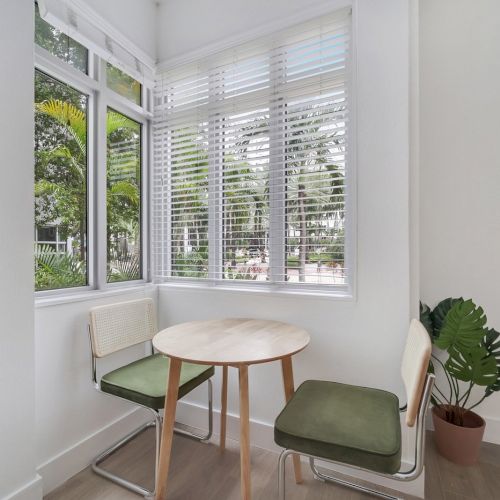 A bright corner nook with a round wooden table, cushioned chairs, potted plant, and white windows with blinds.