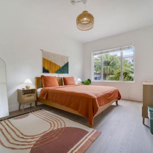 A bright modern bedroom with orange bedding, a patterned rug, bedside tables, palm-view windows, and a wall-mounted TV.