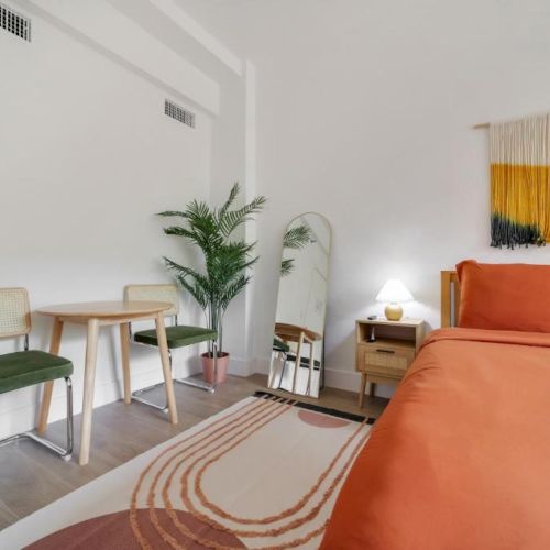 A bright modern living room with an orange sofa, patterned rug, two wooden chairs, a potted plant, lamp, and colorful wall art.