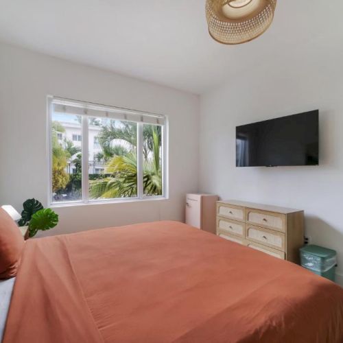 A bright bedroom with a bed, wall-mounted TV, window view of palm trees, and a woven light fixture overhead.