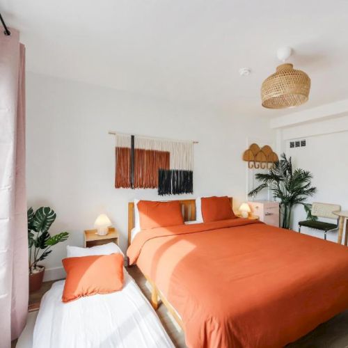 A bright modern bedroom with orange bedding, plants, a woven pendant light, and abstract wall art.