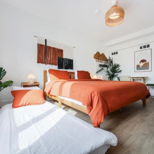 A bright modern bedroom with two beds dressed in orange and white linens, plants, wall art, and a woven pendant lamp overhead.