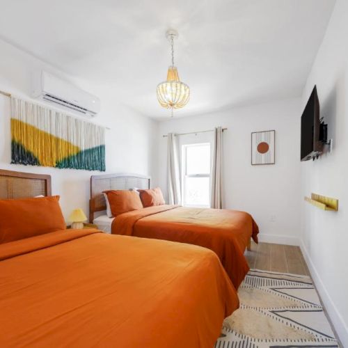 Bright bedroom with three orange beds, pendant light, TV on wall, curtains, and wall art.
