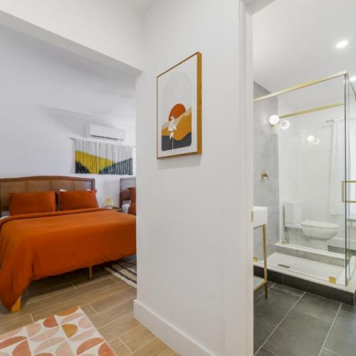 A modern bedroom with an orange bed and an ensuite bathroom visible through a glass wall, lit by ceiling lights.