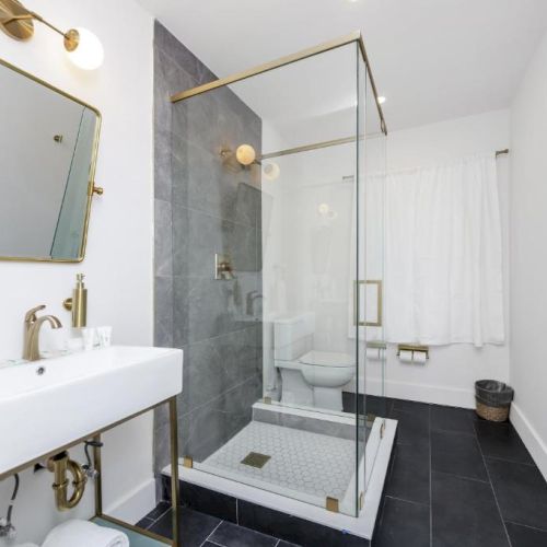 Bright modern bathroom with a white sink, gold mirror, and glass shower with dark gray and gray tile flooring.