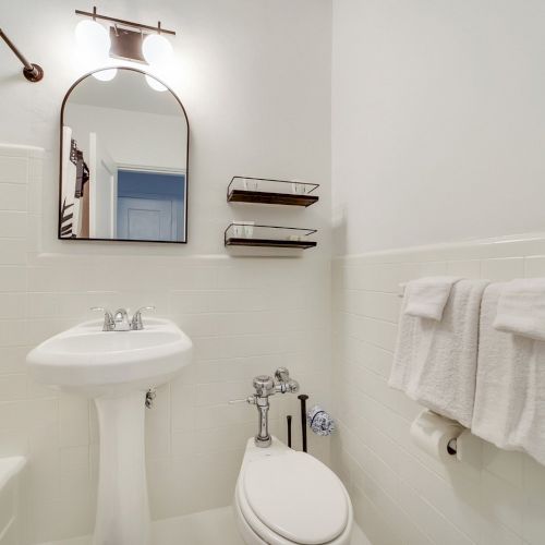 A clean white bathroom with a sink, arched mirror, glass shelves, towel racks, and a toilet.