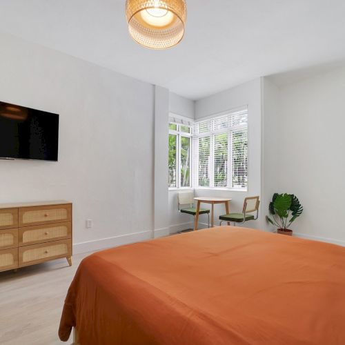 A bright modern bedroom with an orange bedspread, wall TV, wood dresser, large window with blinds, chairs, pendant light, and a potted plant.