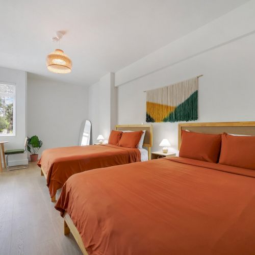 A modern bright bedroom with two beds, orange bedding, a woven pendant light, large window, and a green-and-orange wall tapestry.