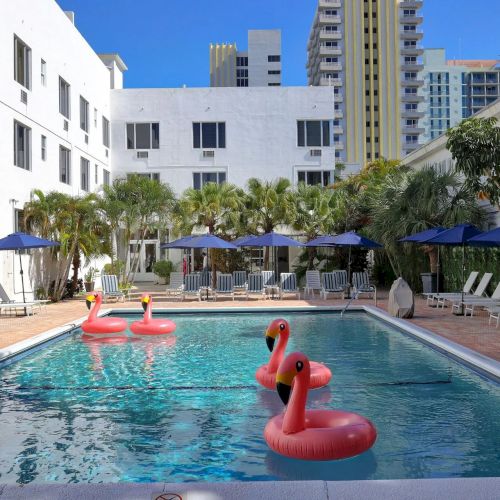 Sunny hotel courtyard pool with inflatable flamingos, blue umbrellas, lounge chairs, and palm trees against tall buildings.