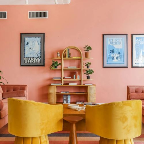 A cozy lounge with peach walls, framed Art Deco posters, a gold bookshelf with plants, and velvet orange/yellow chairs and sofas.
