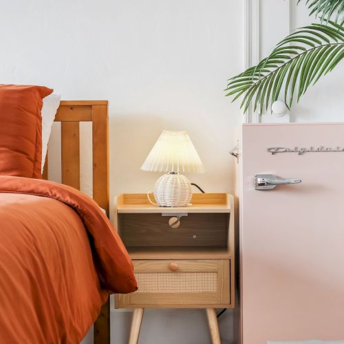 A cozy bedroom corner with orange bedding, a wooden bedside table, a white lamp, and potted tropical leaves.