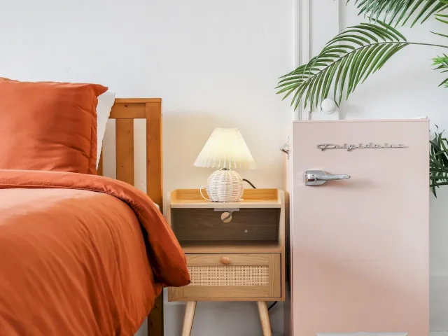 A guest room with an orange bed, wooden nightstand, pink fridge, and white lamp.