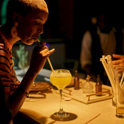 A woman at a dim restaurant table eats with chopsticks, sipping a drink in warm neon lighting while others sit nearby.