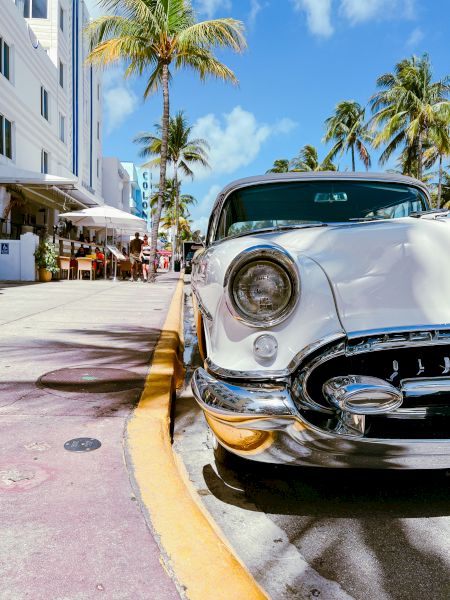 A white classic car sits by a hotel walkway with palm trees and caf&eacute; tables under a bright blue sky.