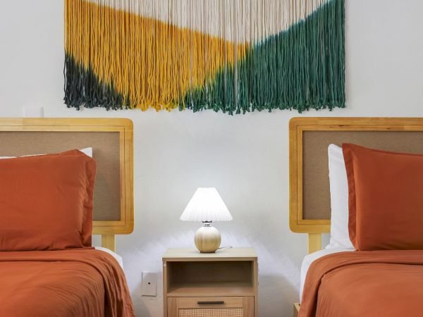A bright twin room with two wooden beds, orange pillows, a bedside lamp, and a striped multicolor wall fringe hanging.