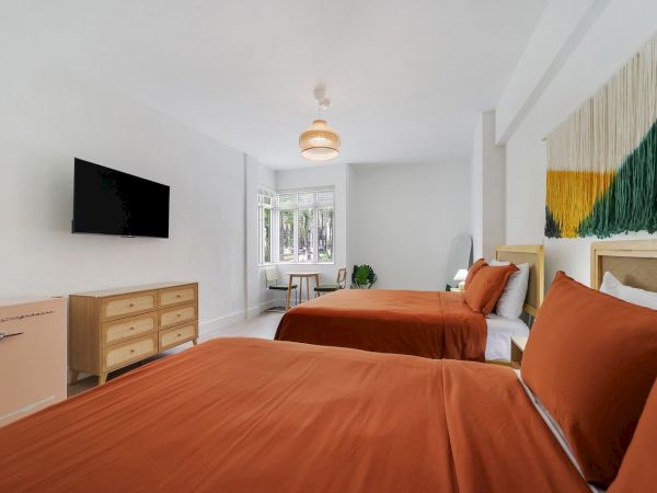 A bright modern hotel bedroom with two beds, orange linens, a TV, and a woven pendant light above.