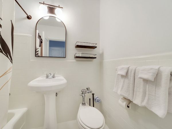 A bright modern bathroom with a pedestal sink, toilet, white tiles, towels, and a wall mirror with overhead lights.
