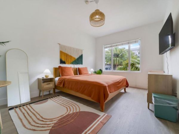 A bright, modern bedroom with an orange bed, patterned rug, bedside tables, palm-window view, and a wall-mounted TV.