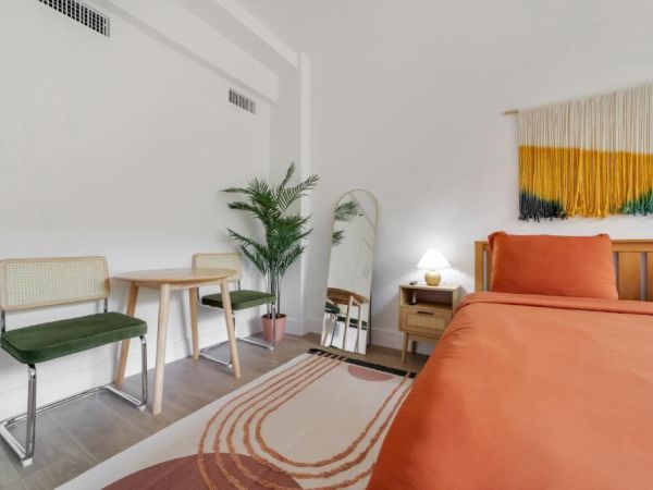 A bright modern living room with an orange sofa, rug, chairs, side table, lamp, and a colorful wall hanging.