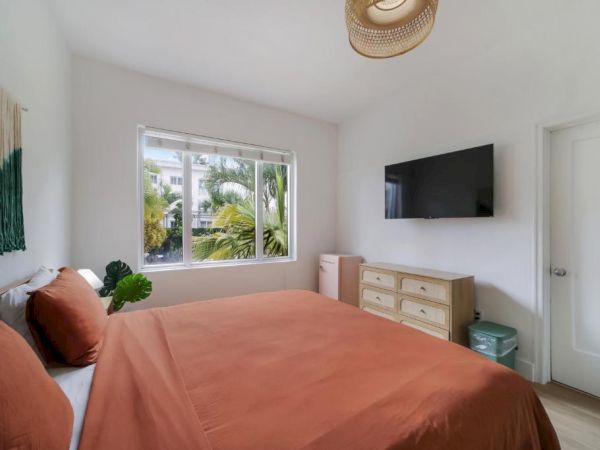 A bright bedroom with a made bed, wall TV, dresser, big window, tropical plants, and a woven ceiling light.